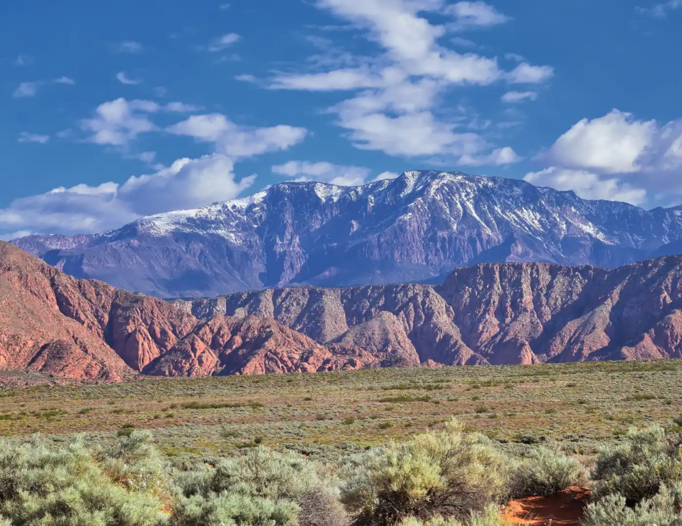 Snow-dusted mountain ridge rising behind banded red sandstone foothills with sagebrush flats in the foreground under a partly cloudy sky
