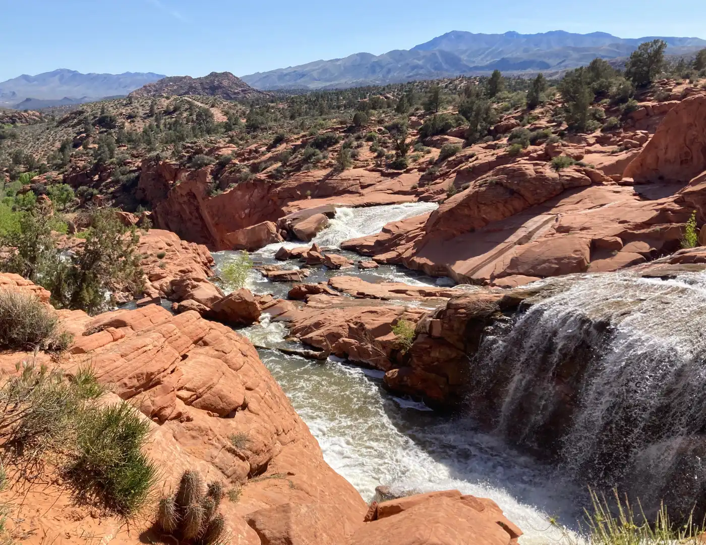 Waterfall cascading over smooth red sandstone with rushing white water, desert vegetation, and distant blue mountains under a clear sky