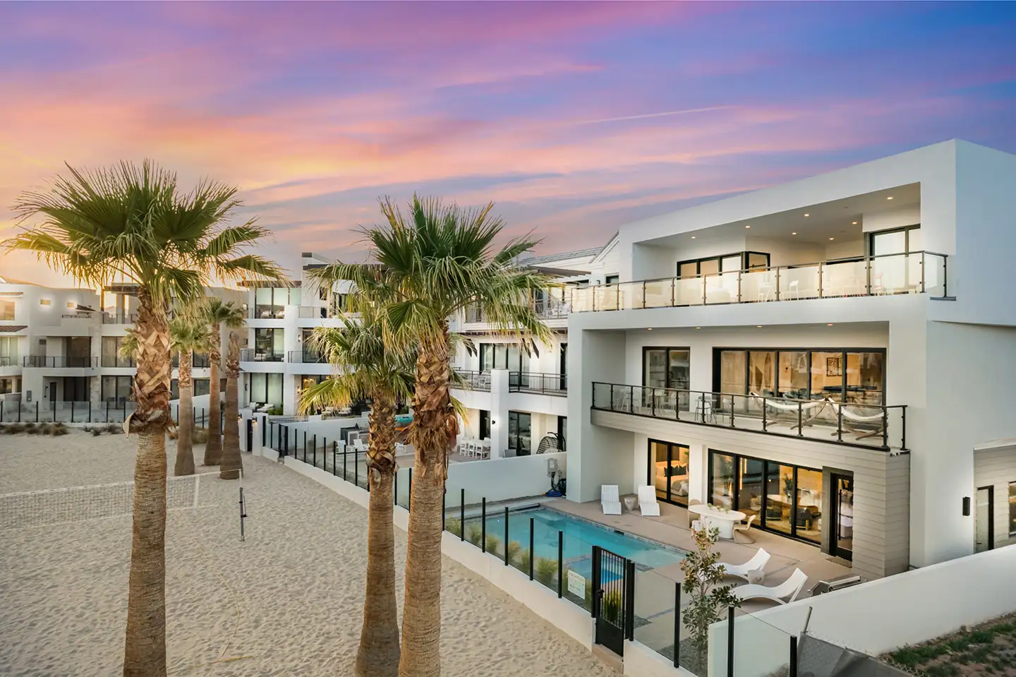 Modern white luxury home with a pool and palm trees beside a sandy area at sunset.