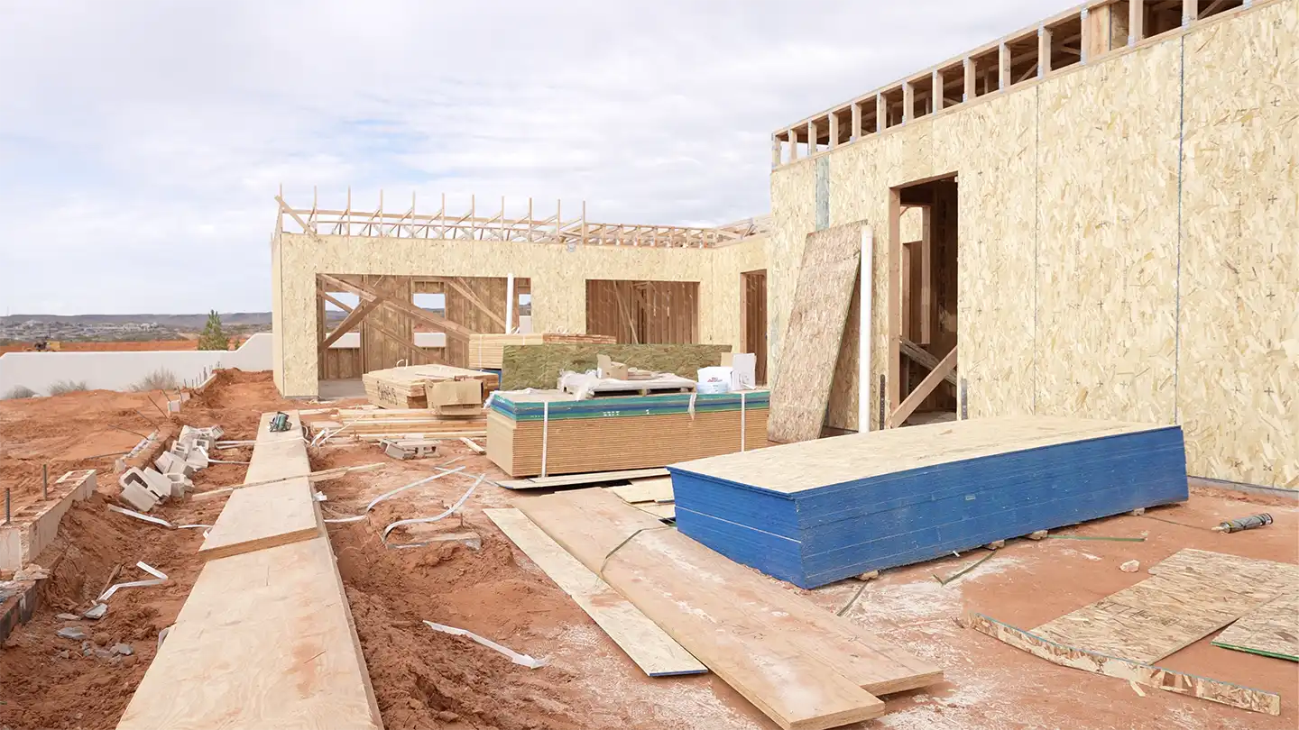Custom home under construction with framed walls and stacked lumber and materials on a desert lot.