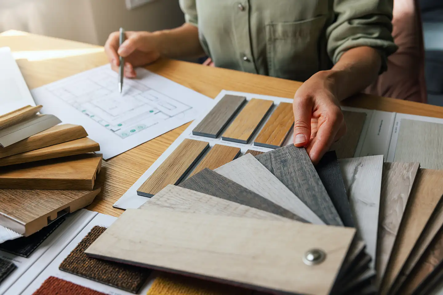 Hands reviewing wood flooring samples and finish materials over a floor plan during a home design selection meeting.