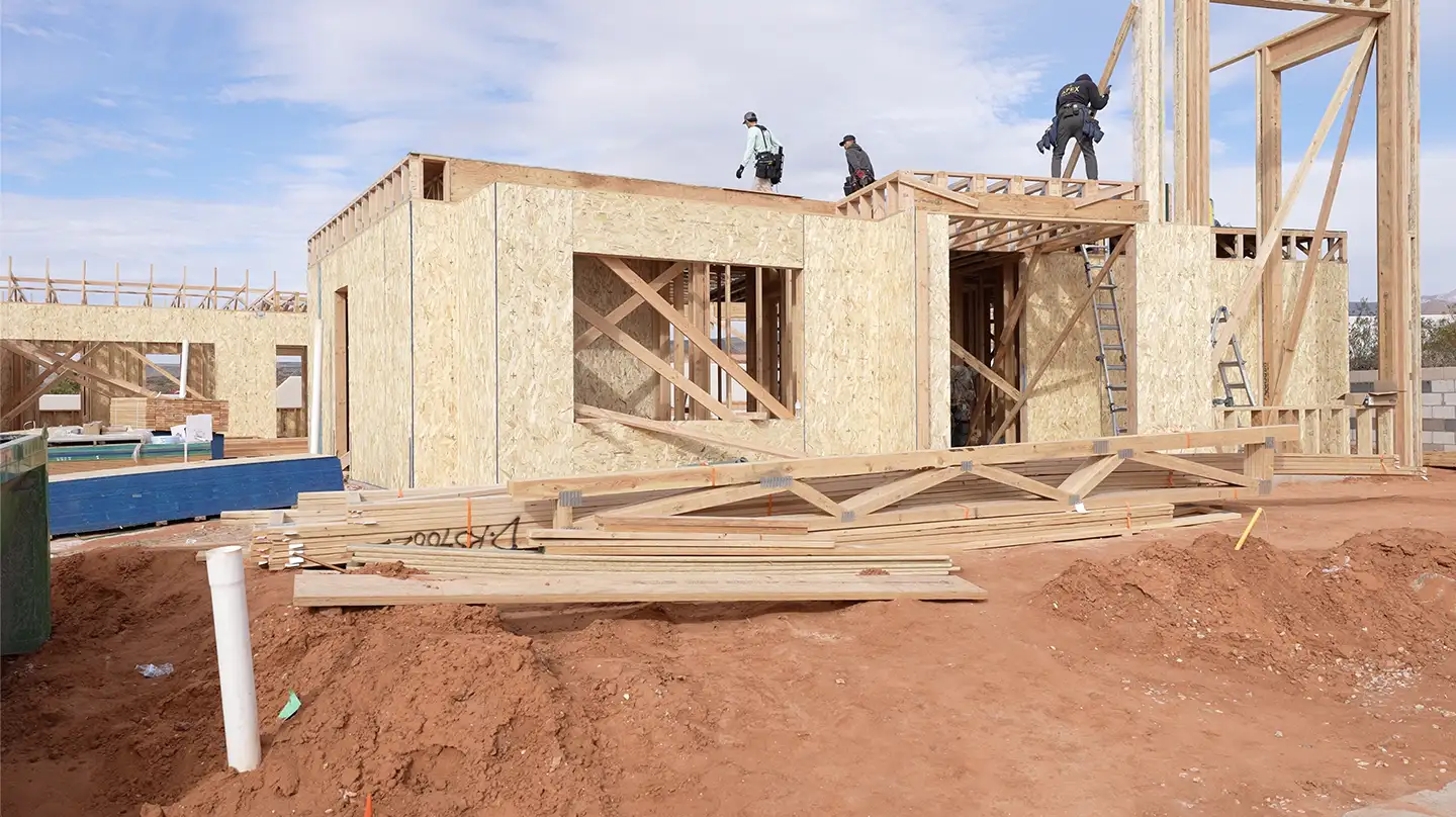 Workers framing a new custom home with wood studs and OSB sheathing on a red-dirt construction site under a blue sky.