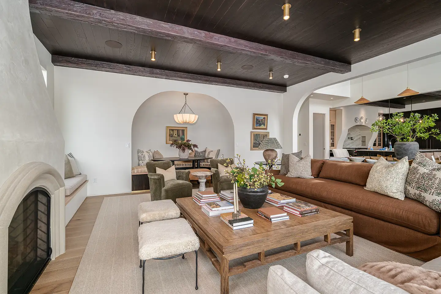 Warm living room with a brown sectional sofa, stucco fireplace, wood-beamed ceiling and arched doorways to a dining area.