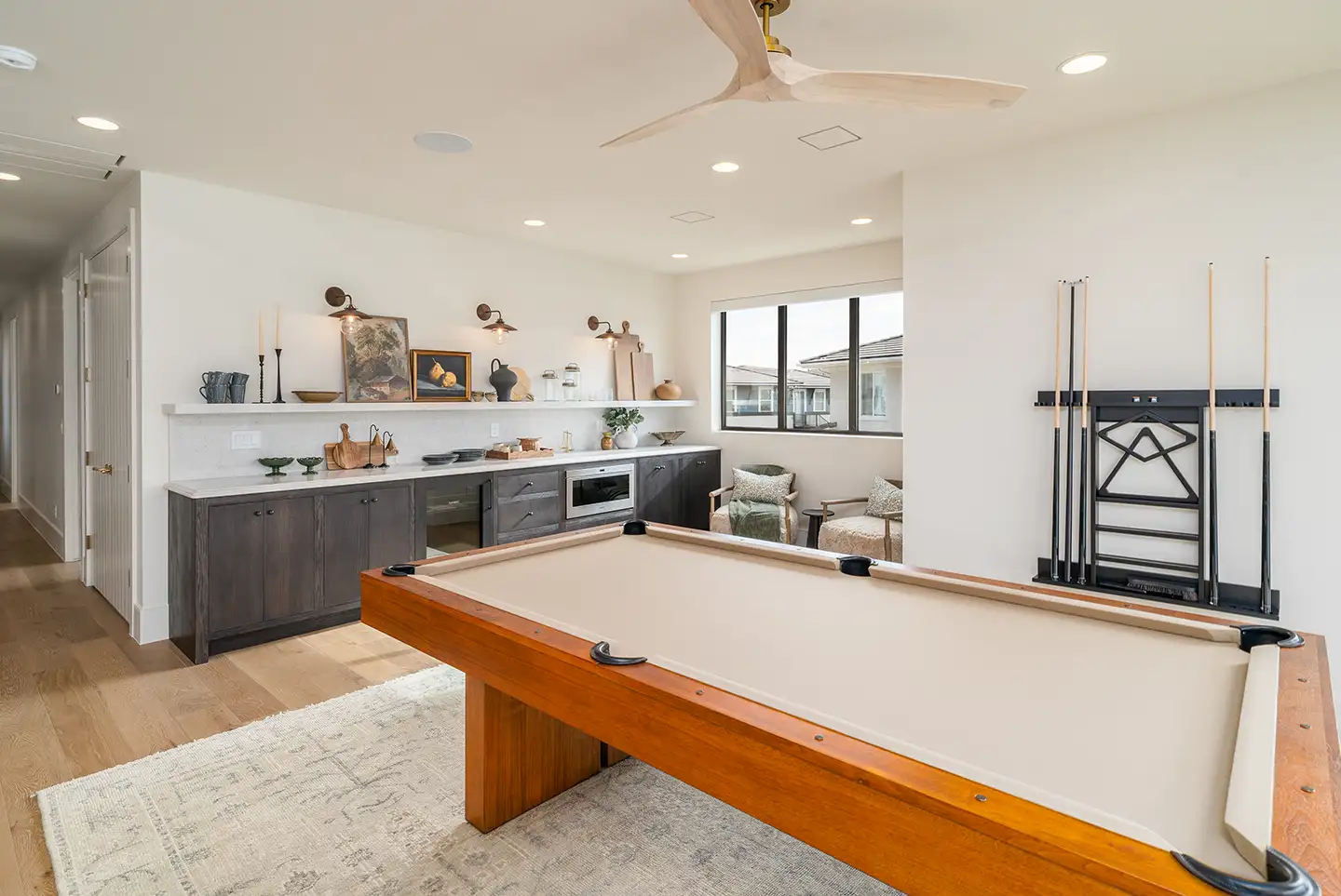 Game room with a wood billiards table, cue rack, bar cabinetry and a seating area beside a window in a luxury home.