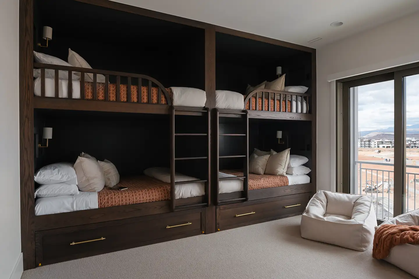 Bunk room with two sets of built-in dark wood bunk beds, sconce lighting and storage drawers beside a window with desert views.
