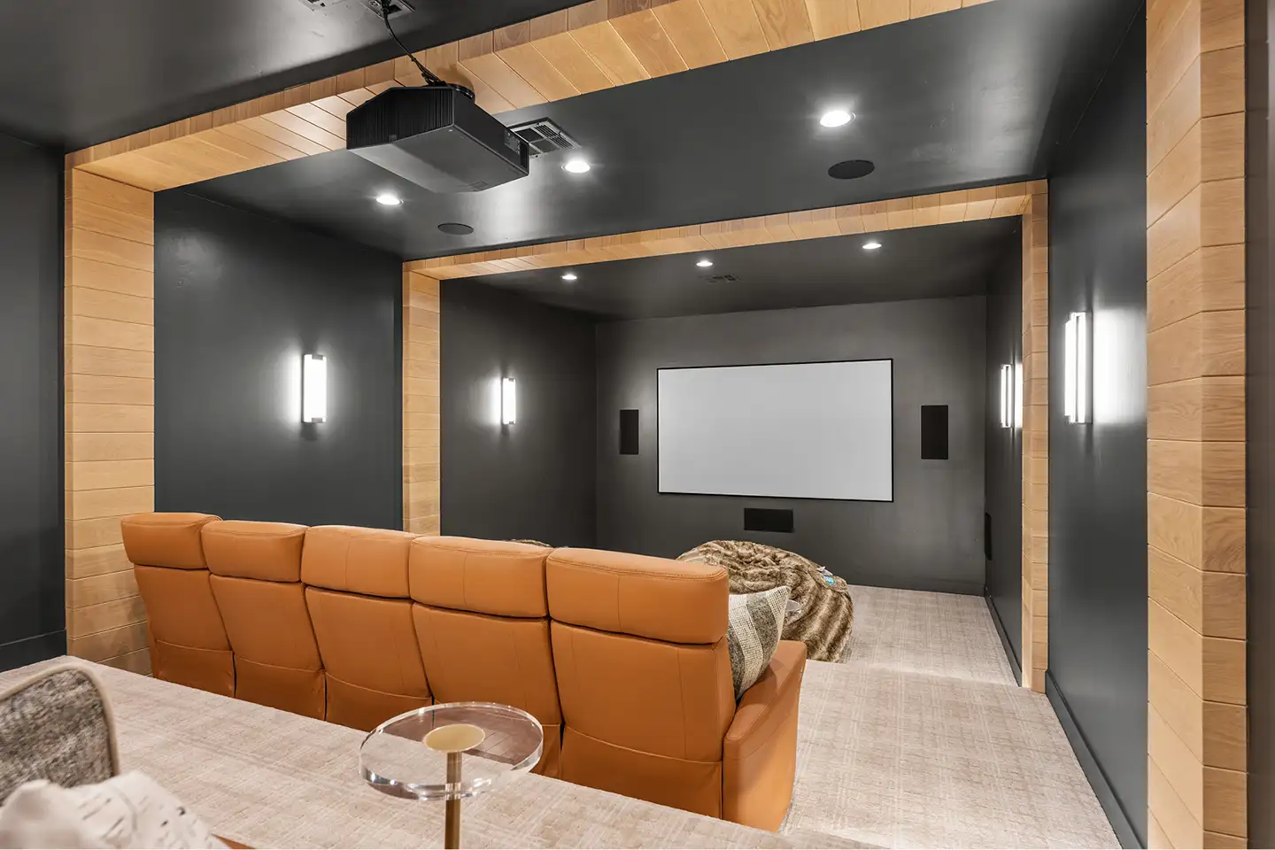Home theater with tan leather reclining seats, a projector and large screen, dark walls and wood-trimmed accent lighting.