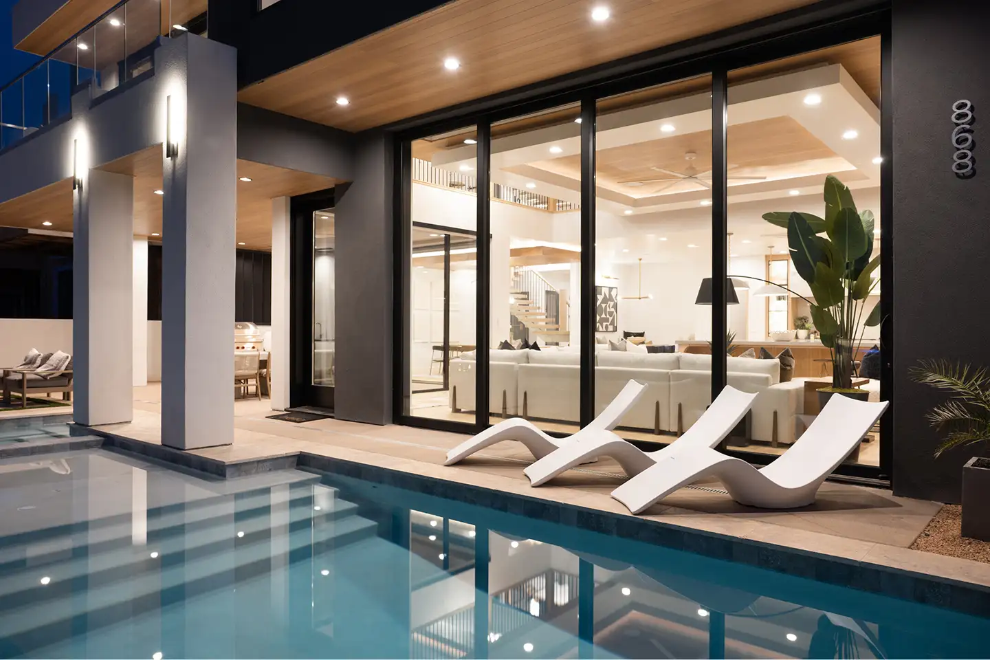 Modern home lit at dusk with a pool, lounge chairs and glass walls revealing the living spaces inside.