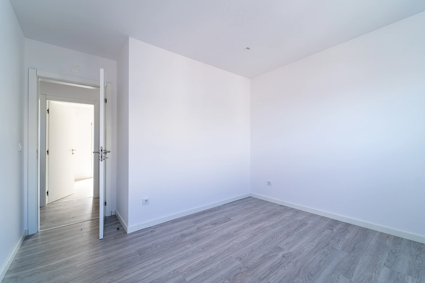 Empty bedroom with plain white walls, gray laminate flooring and an open door to a hallway in a newly built home.