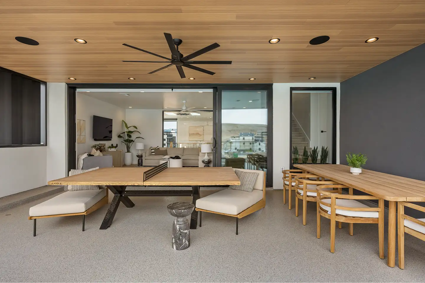 Covered deck with an outdoor ping-pong table, dining seating and a wood-plank ceiling, opening to the living room through glass sliders.