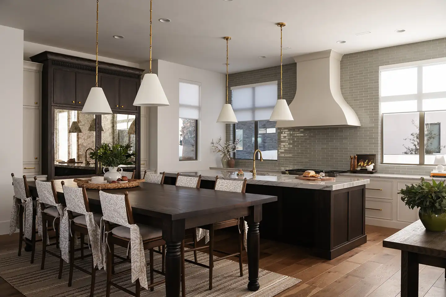 Luxury kitchen with a large marble island, white plaster range hood, gray tile backsplash, brass pendants and a dark-wood dining table.