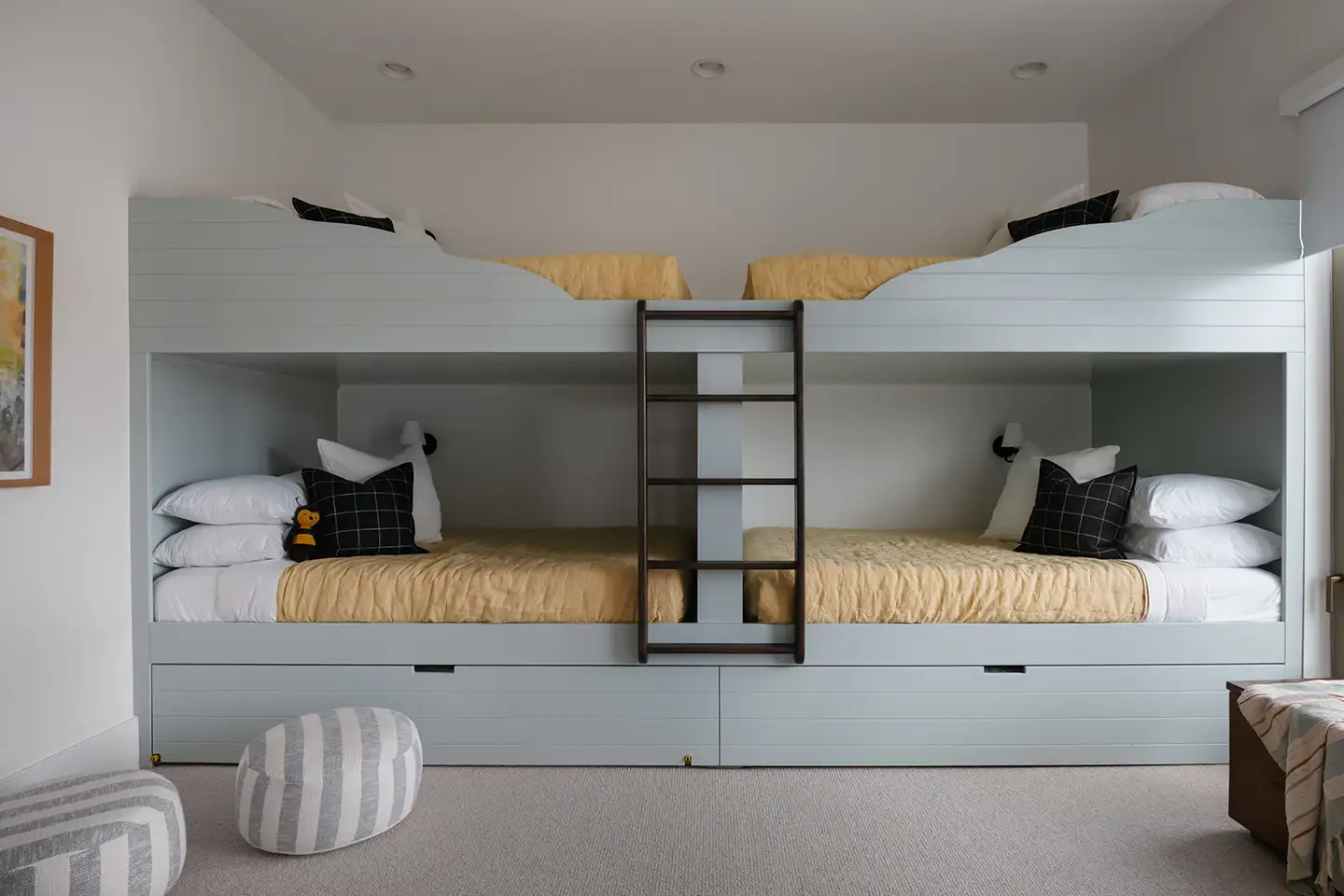 Bunk room with two built-in double bunk beds in pale-blue millwork, a center ladder, yellow bedding and striped floor poufs.