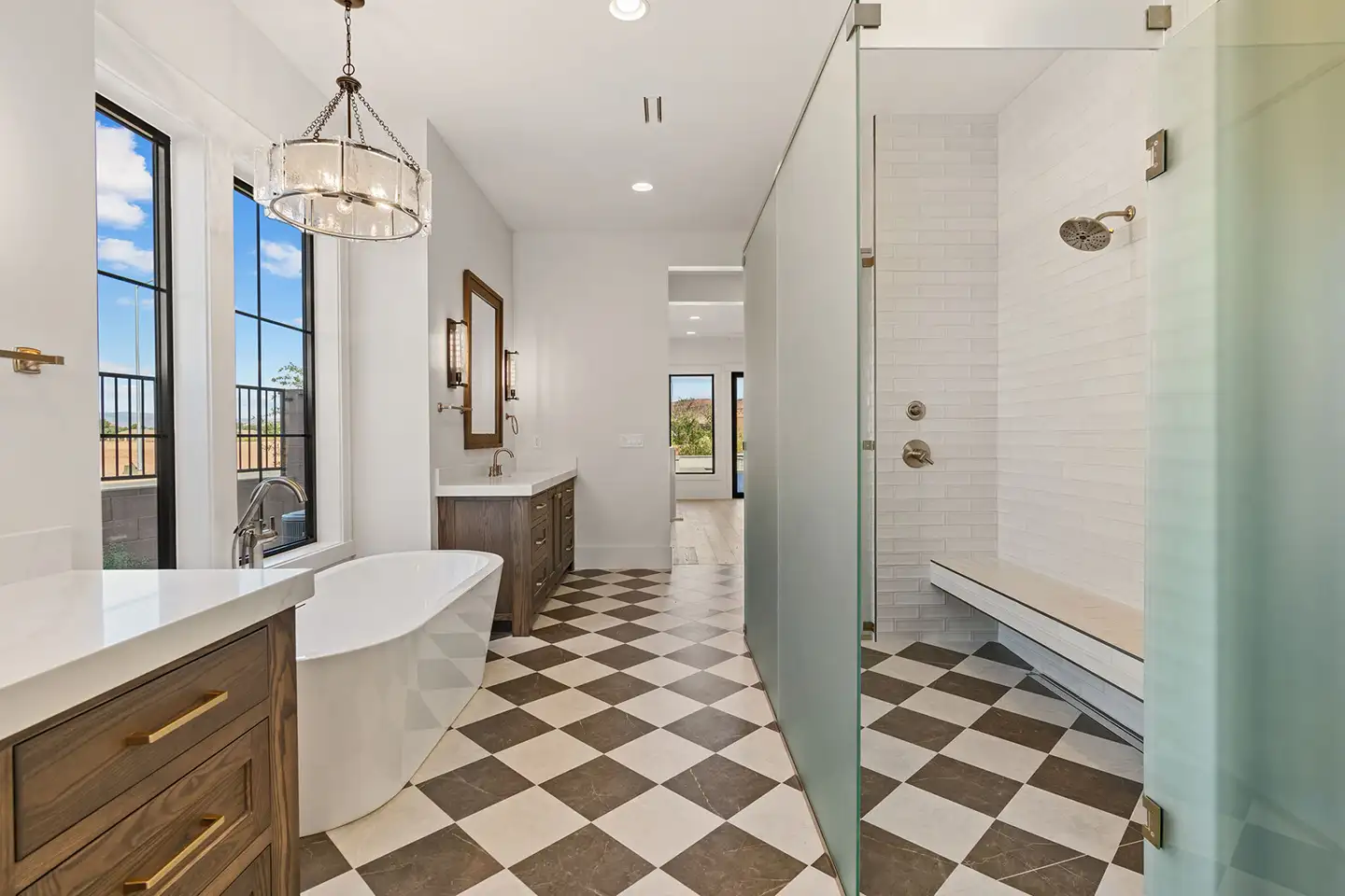 Luxury primary bathroom with a freestanding soaking tub, checkerboard tile floor, walk-in glass shower and dual wood vanities.