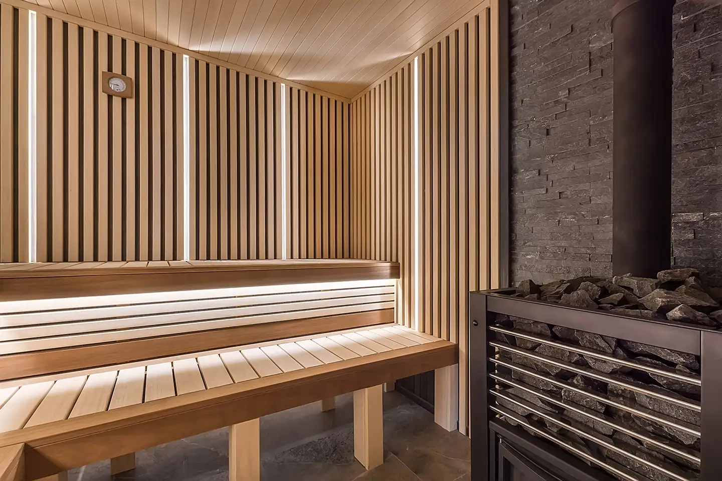 Infrared sauna with backlit wood-slat walls, tiered bench seating and a stone heater filled with sauna rocks.