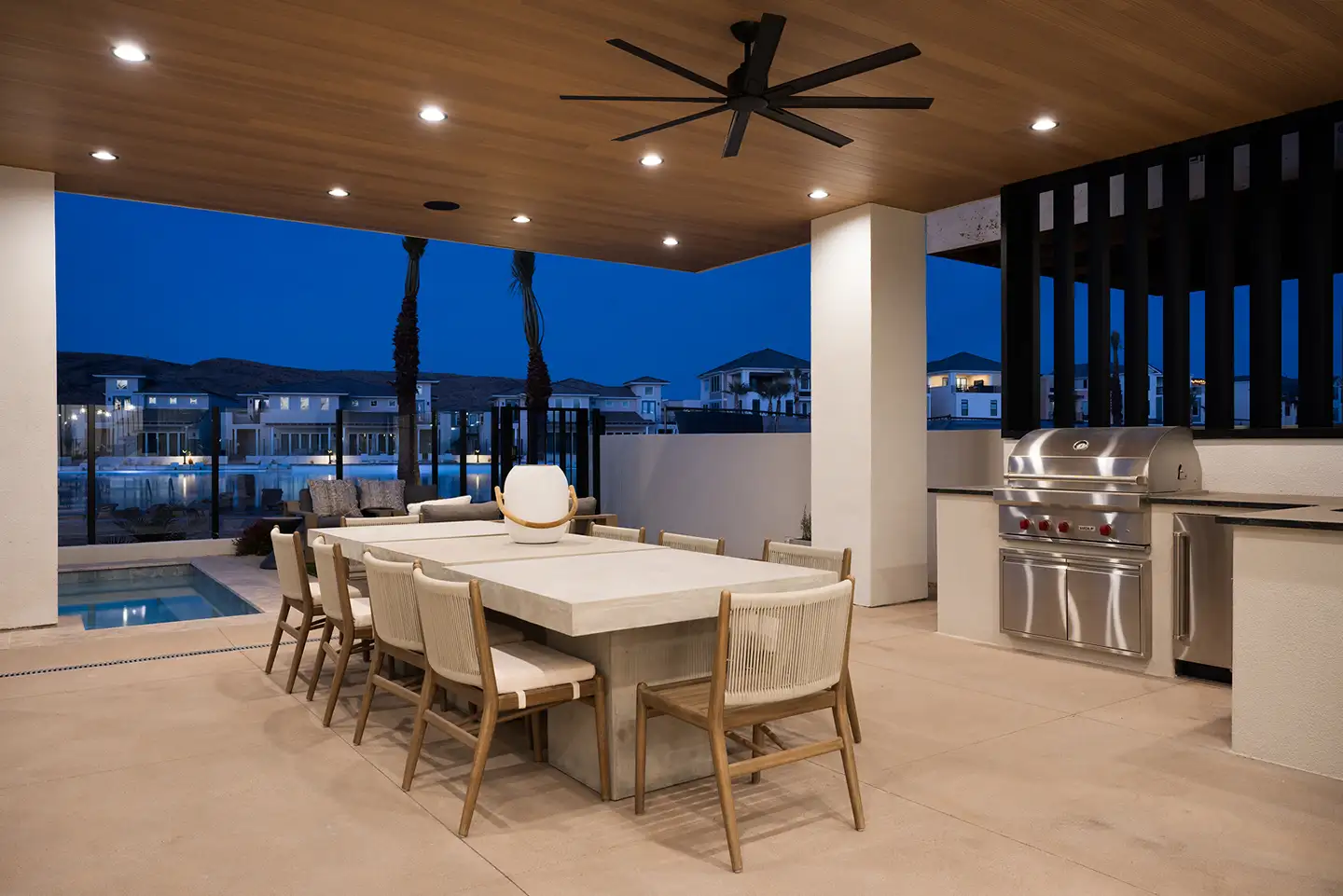 Covered patio at dusk with a concrete dining table, large ceiling fan, built-in stainless grill and a pool with community views.