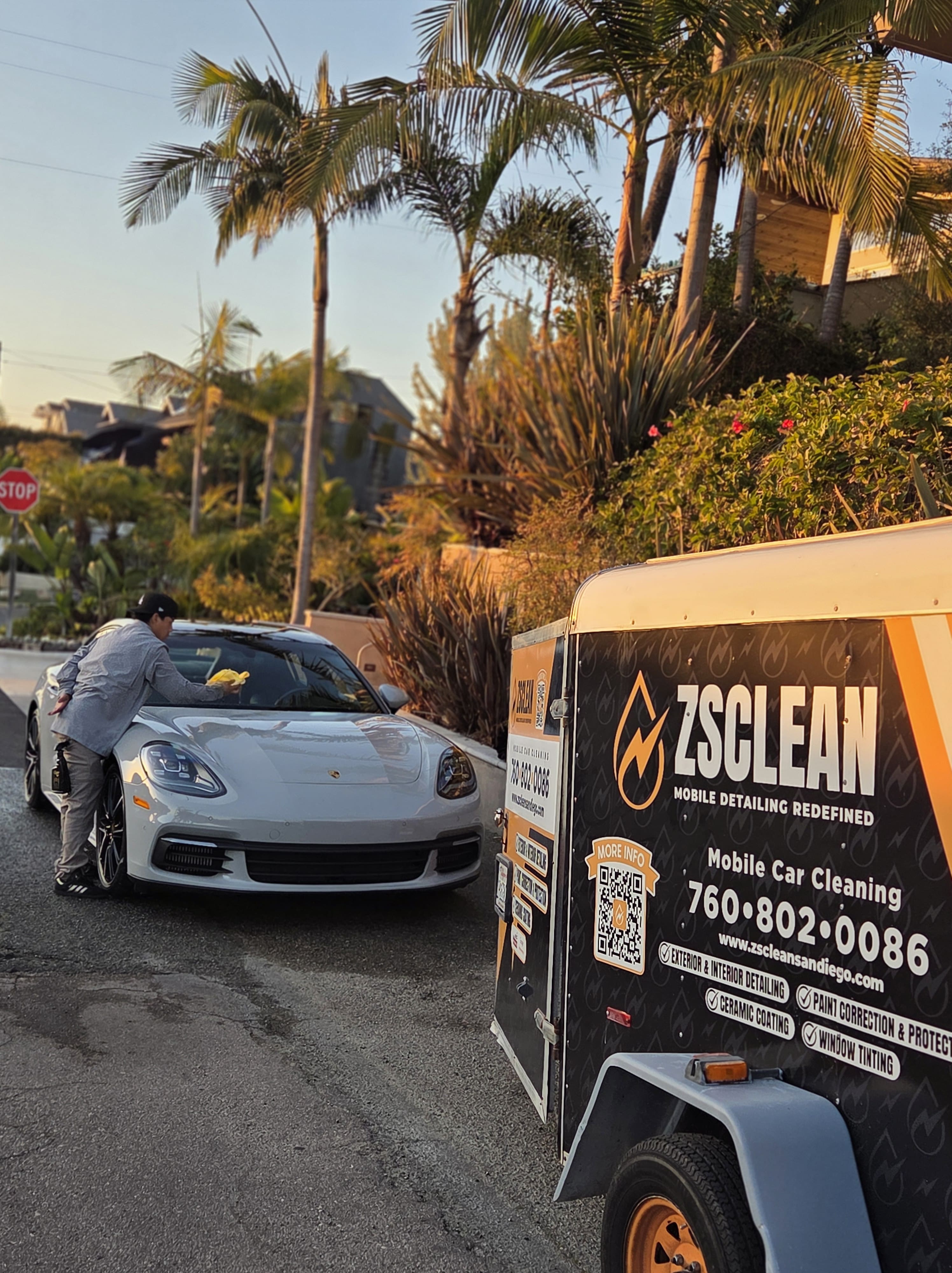 Person cleaning the hood of a white Porsche car parked on a street next to a ZSCLEAN mobile car cleaning trailer.