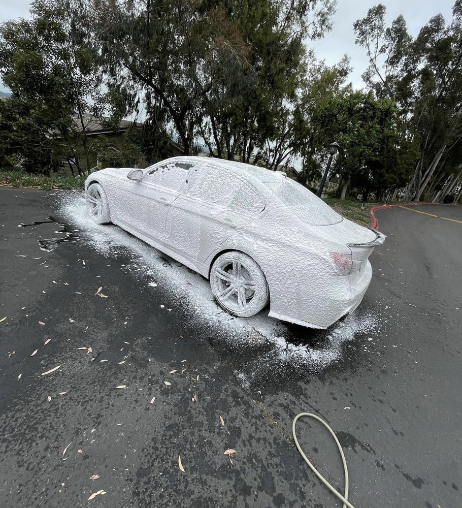 White car covered in thick foam during a wash, parked on asphalt with trees in the background.