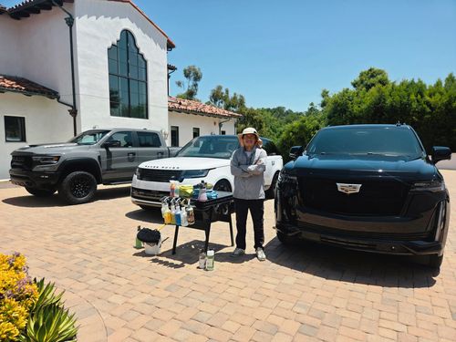 Person standing with arms crossed beside three parked SUVs and a cart full of car cleaning supplies in a sunlit driveway.