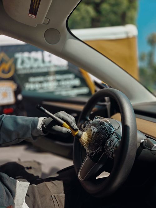 Person wearing gloves cleaning a car steering wheel with a small brush and cleaning foam inside a vehicle.