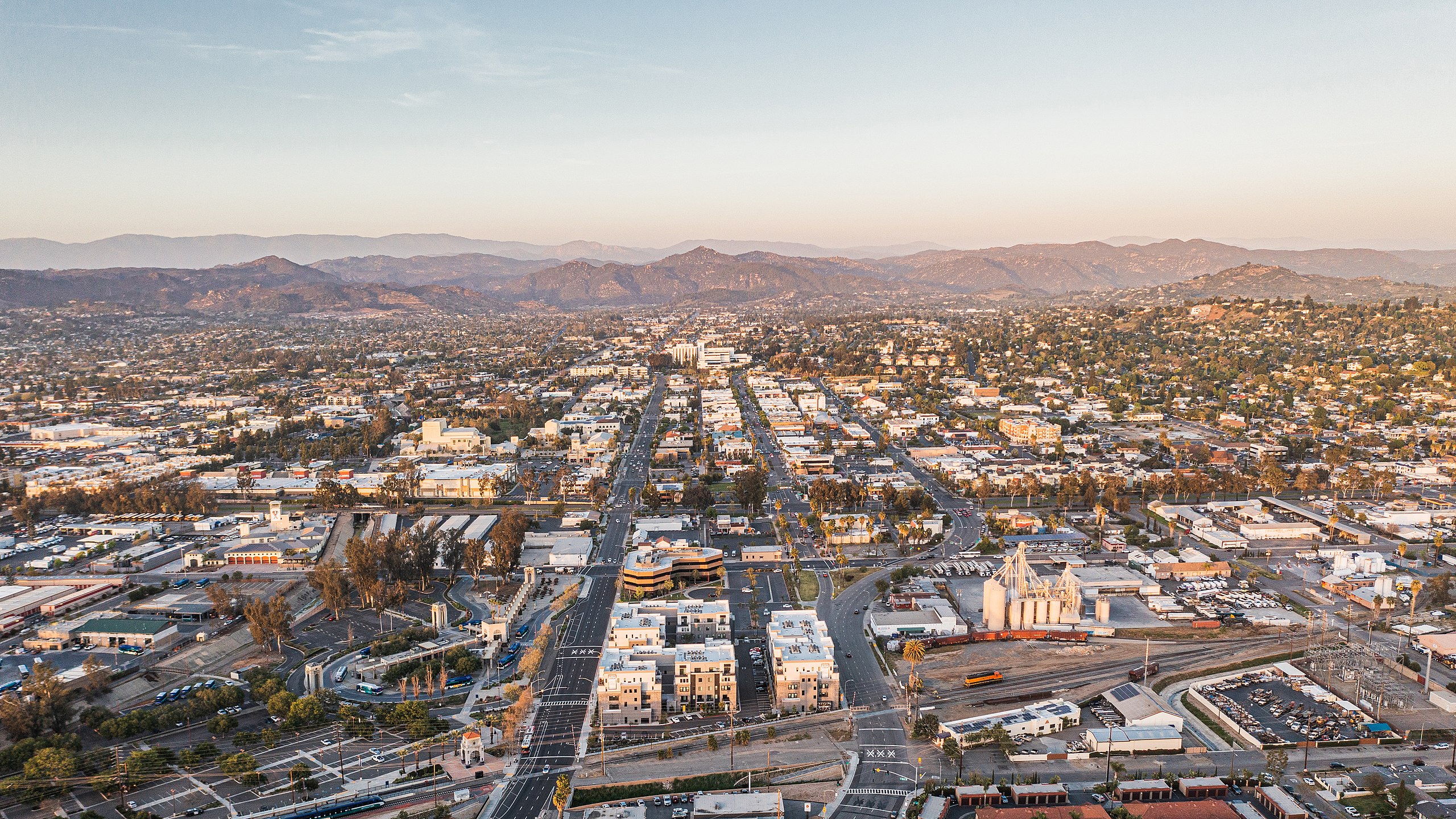 Aerial view of a city with grid streets, buildings, and mountains in the distance under a clear sky.