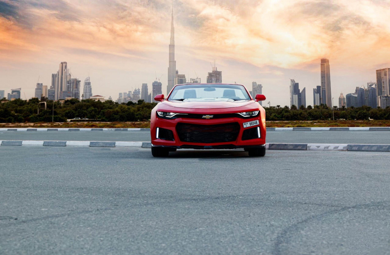 Red Chevrolet sports car parked with Dubai skyline at sunset