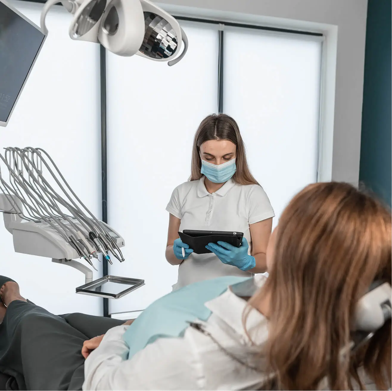 Female dental assistant wearing a mask and gloves reviewing patient information on a tablet while a female patient reclines in the dental chair.