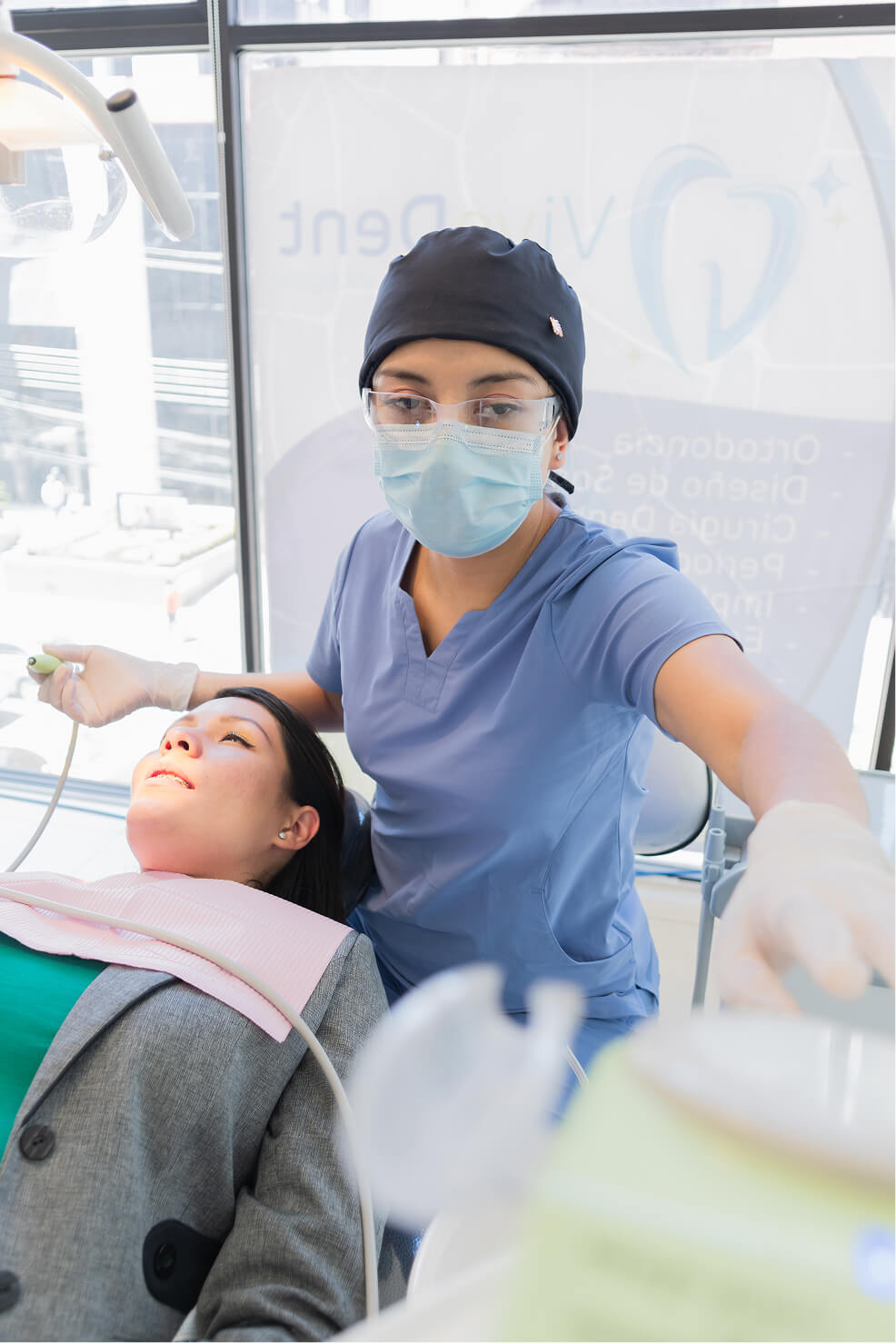 Female dental professional in scrubs and a face mask preparing equipment while a female patient reclines in the dental chair under examination light.