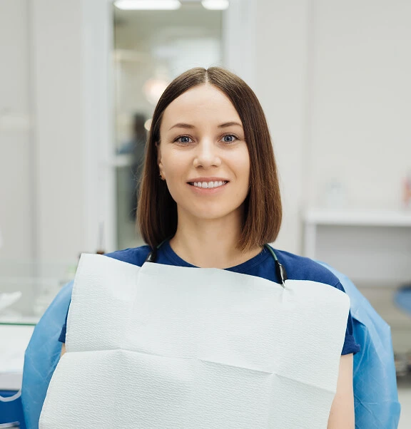 Female dental patient smiling while seated in a dental chair wearing a protective bib, ready for examination or treatment.