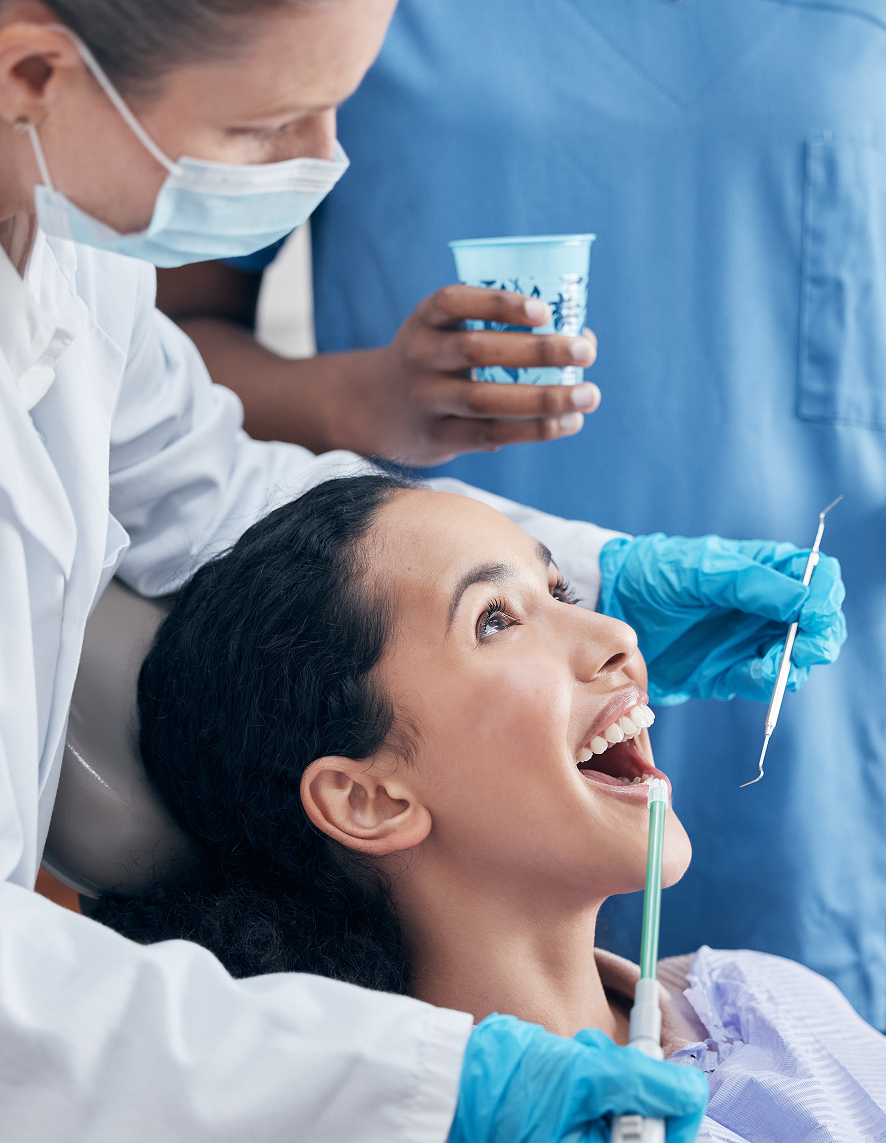 Smiling female patient receiving a dental cleaning from a masked hygienist using suction and scaler tools, with a dental assistant holding a rinsing cup in the background.
