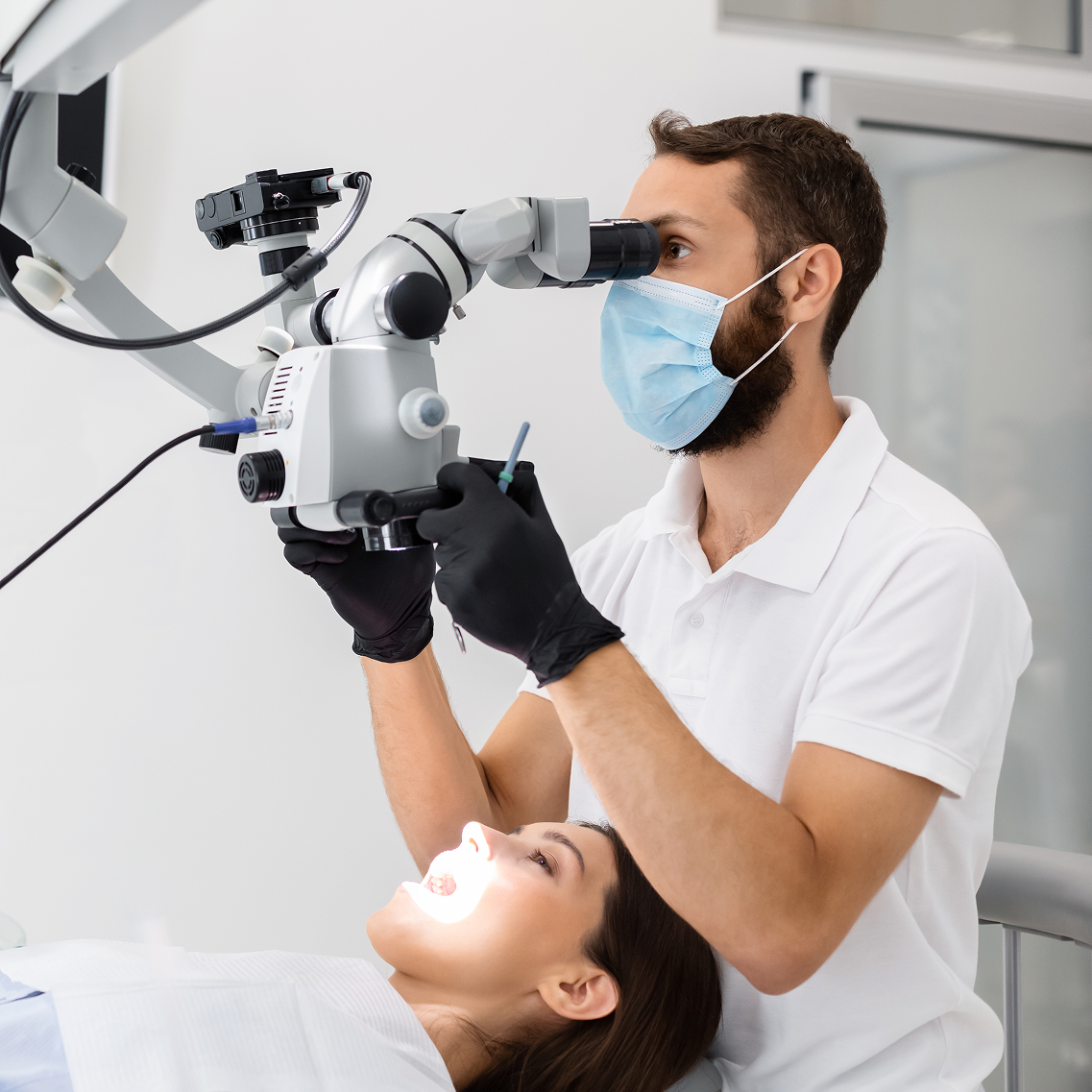 Dentist wearing a mask and gloves using a dental microscope to perform a precision procedure on a female patient with mouth open under examination light.