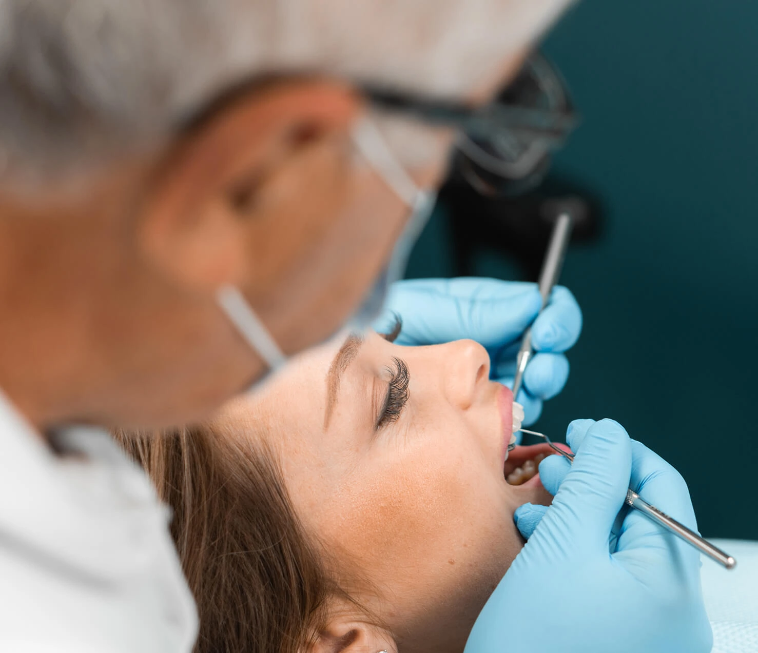 Close-up of a female patient receiving a dental exam from a masked dentist using a mouth mirror and explorer tool while wearing blue gloves.