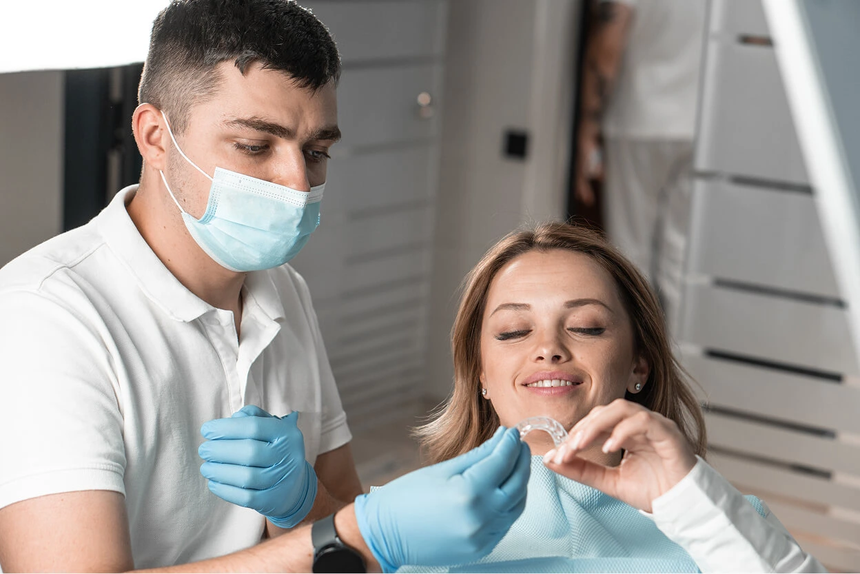 Female dental patient holding a clear aligner while seated in a dental chair, with a masked dentist wearing gloves guiding her during an orthodontic consultation.
