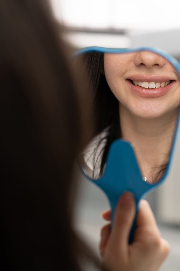 Close-up of a female dental patient smiling at her reflection in a handheld blue mirror after treatment.
