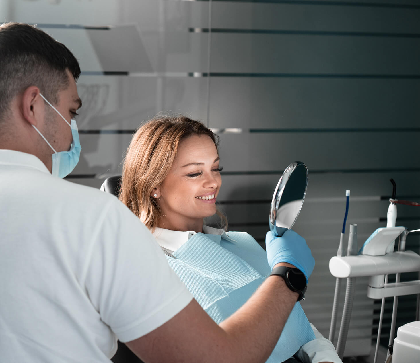 Female patient smiling while looking into a mirror during a dental checkup with a masked dentist in a modern clinic.