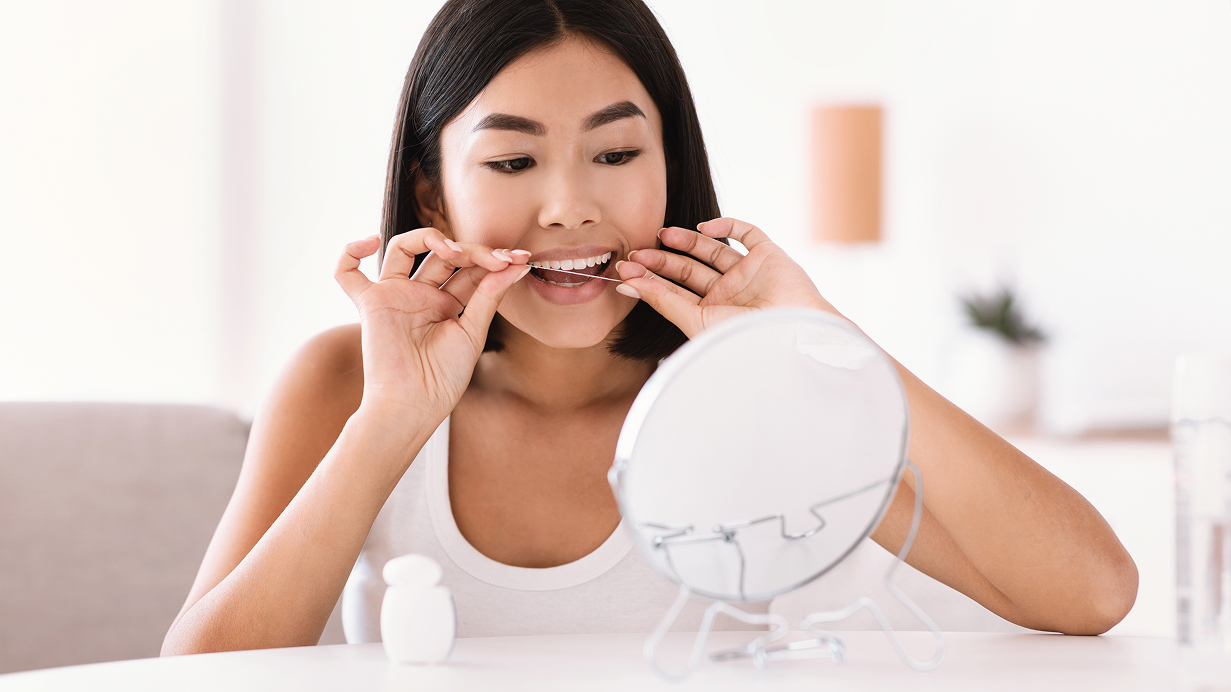 Young woman using dental floss in front of a bathroom mirror to clean between her teeth as part of her oral hygiene routine.