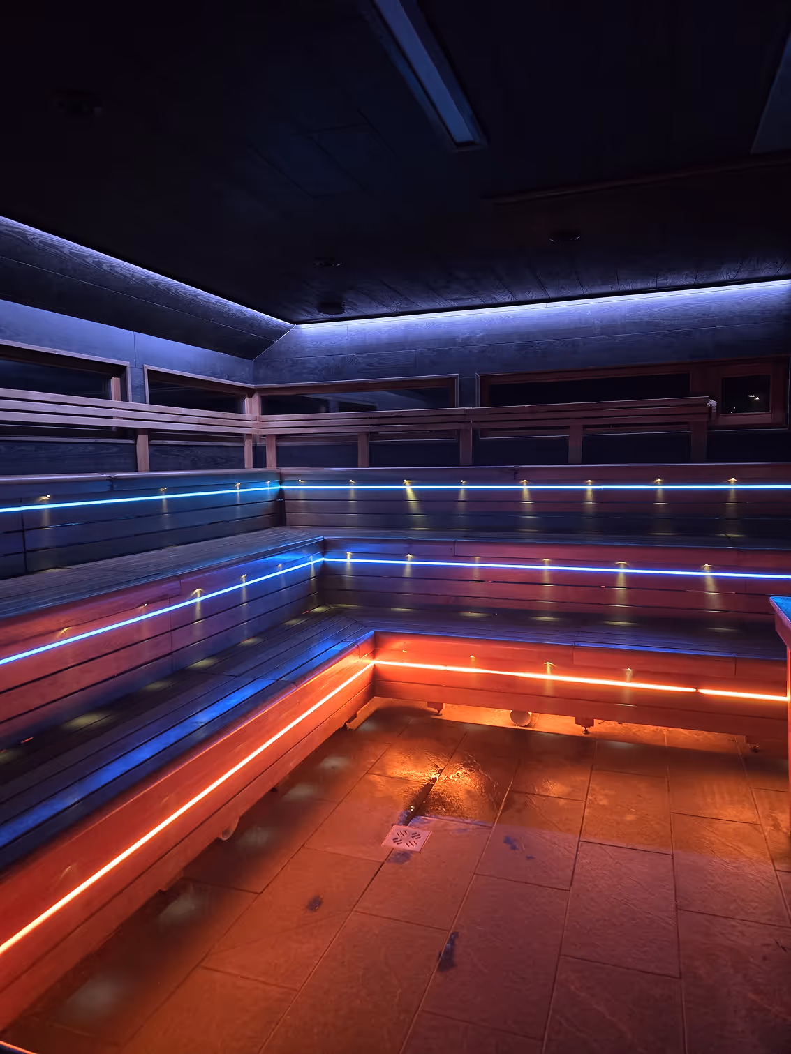Interior of a modern sauna with wooden benches lit by blue and orange LED strip lights.