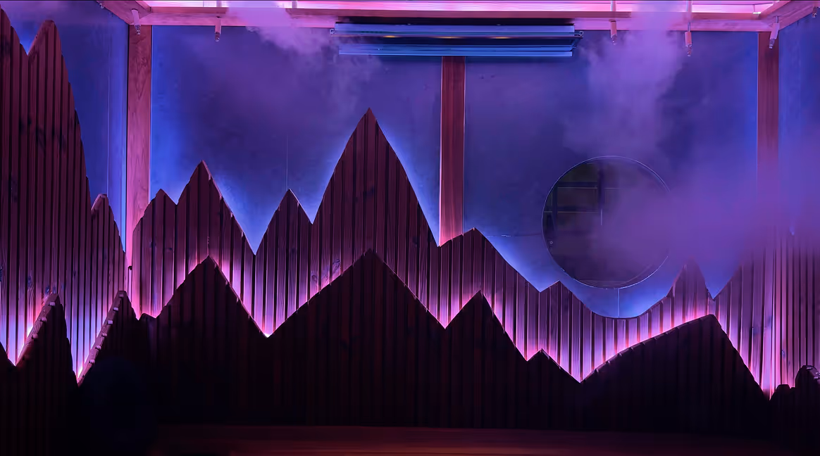 Indoor wooden wall designed with triangular peaks resembling mountain silhouettes, illuminated with purple and blue lighting and surrounded by mist.