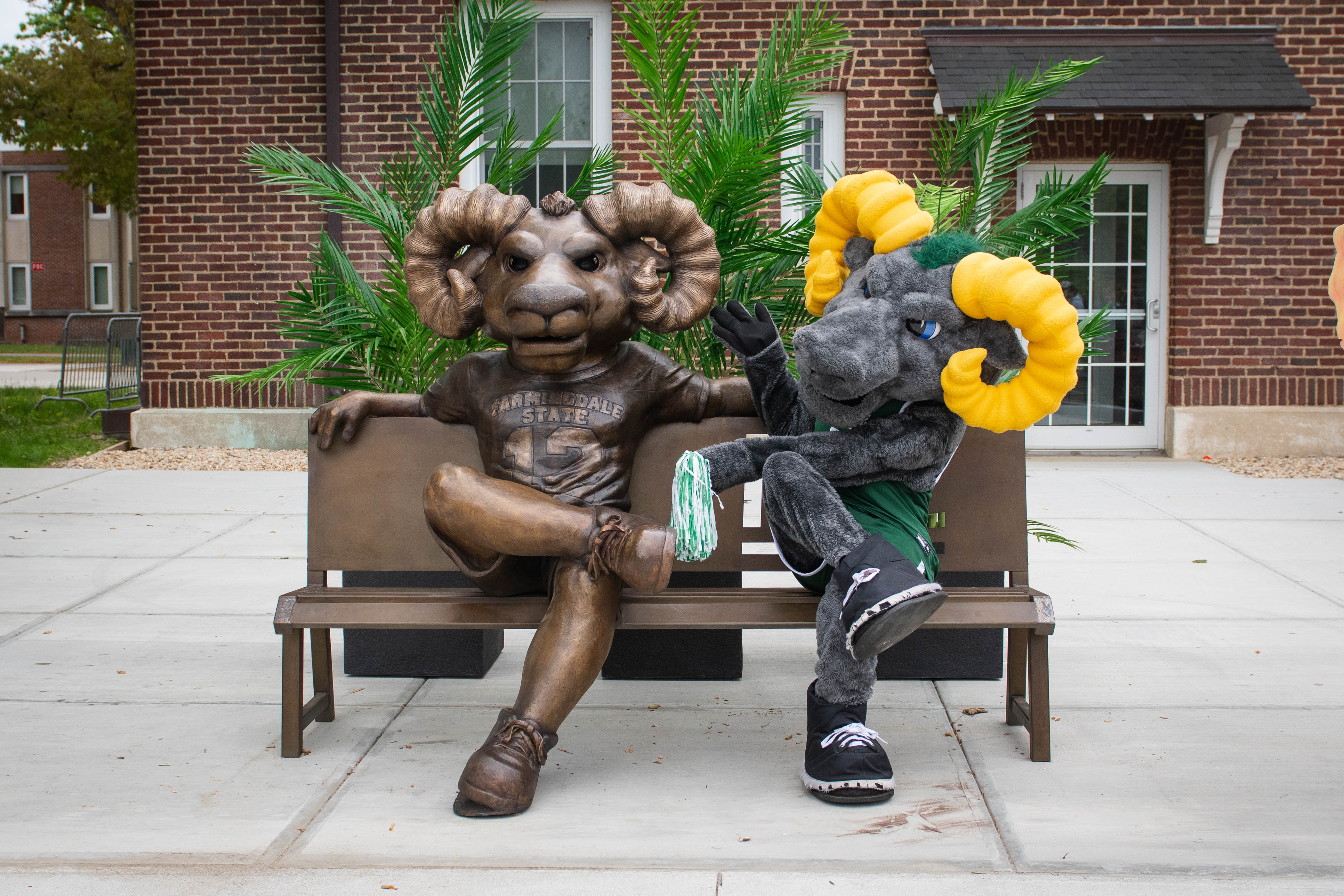 Brown ram statue and gray ram mascot with yellow horns sitting on a bench outside a brick building.