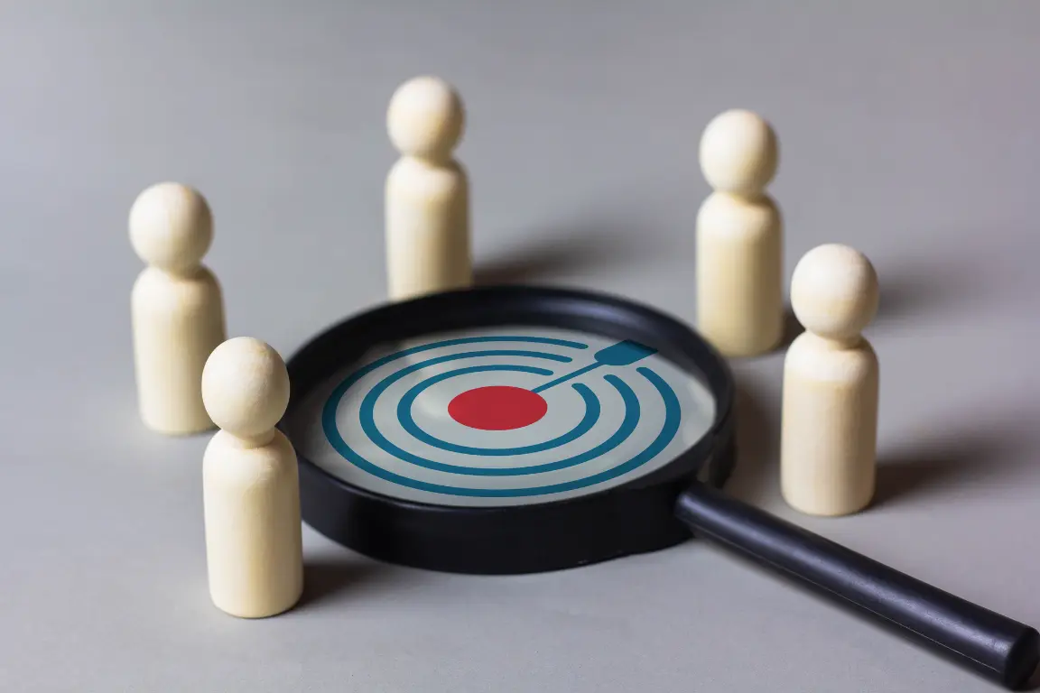 Five wooden figurines surrounding a magnifying glass focusing on a red target bullseye with concentric blue circles.