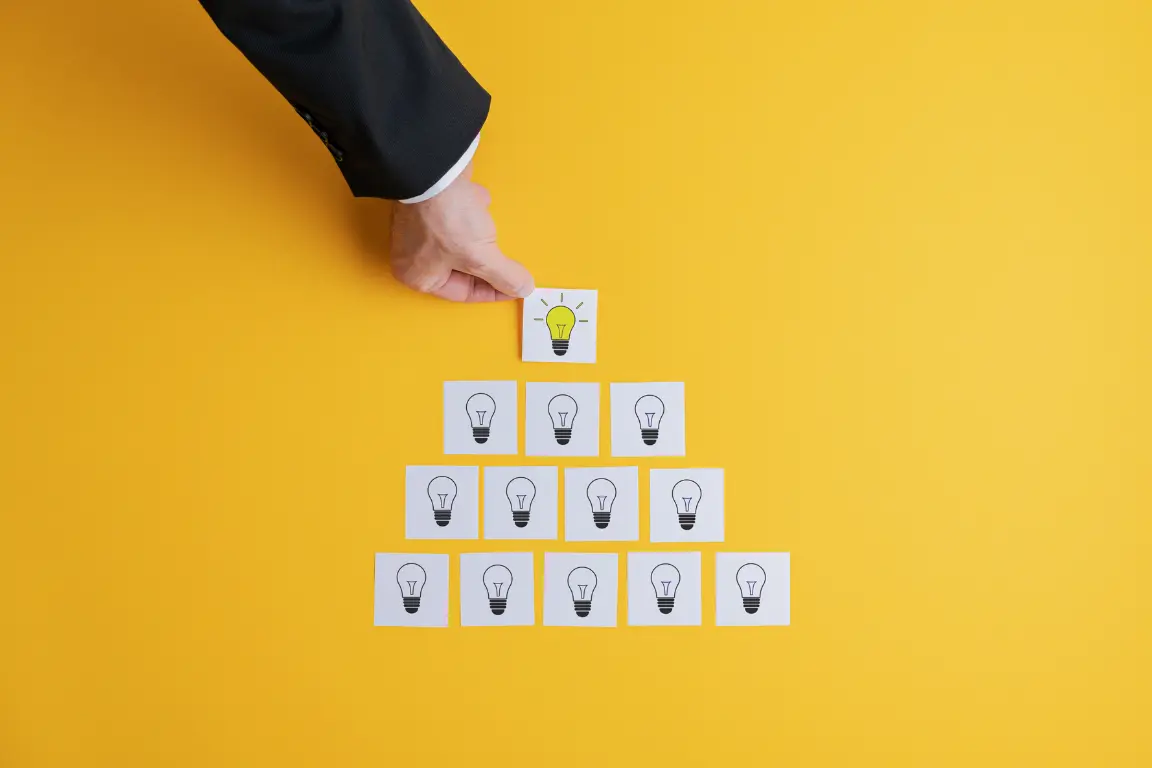 Hand in business suit placing a yellow light bulb card on top of a pyramid of white cards with black light bulb icons on a yellow background.