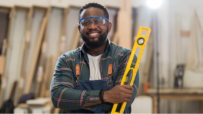 A smiling handyman in a workshop wearing safety goggles and holding a yellow spirit level.
