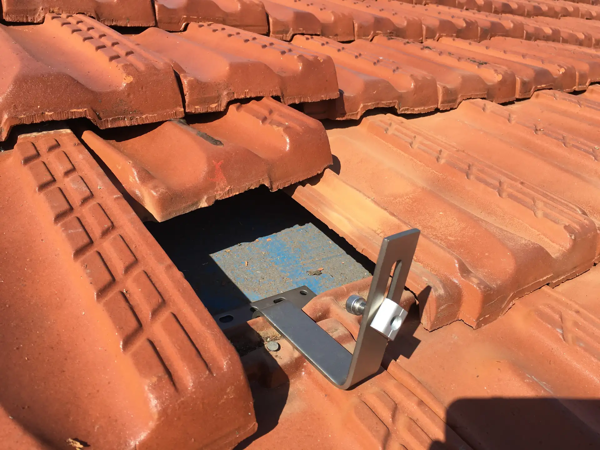 Close-up of a Clenergy roof mounting bracket secured to a rafter beneath a terracotta tile, showing the flashing base plate — Solarbank installation
