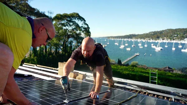 Two Solarbank installers fixing black solar panels to a tiled roof overlooking a Sydney waterway