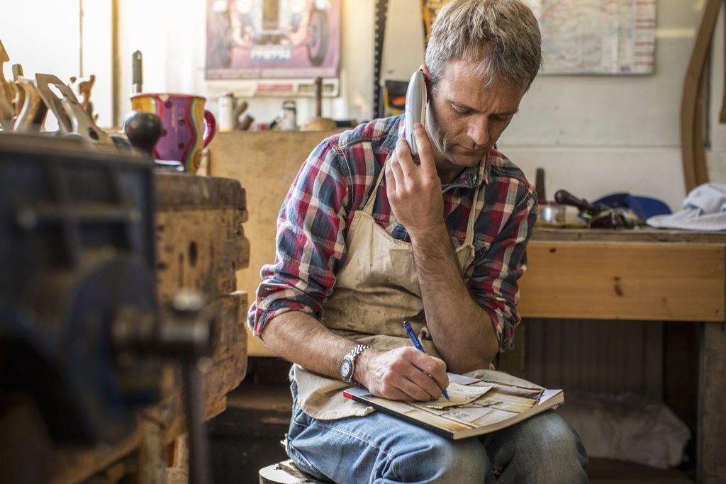 Ein Handwerker sitzt in seiner Werkstatt, klemmt sich ein Telefon ans Ohr und macht sich gleichzeitig konzentriert Notizen in ein Notizbuch. Er trägt ein kariertes Hemd und eine Arbeitsschürze, was die geschäftige Atmosphäre eines Kleinunternehmers unterst