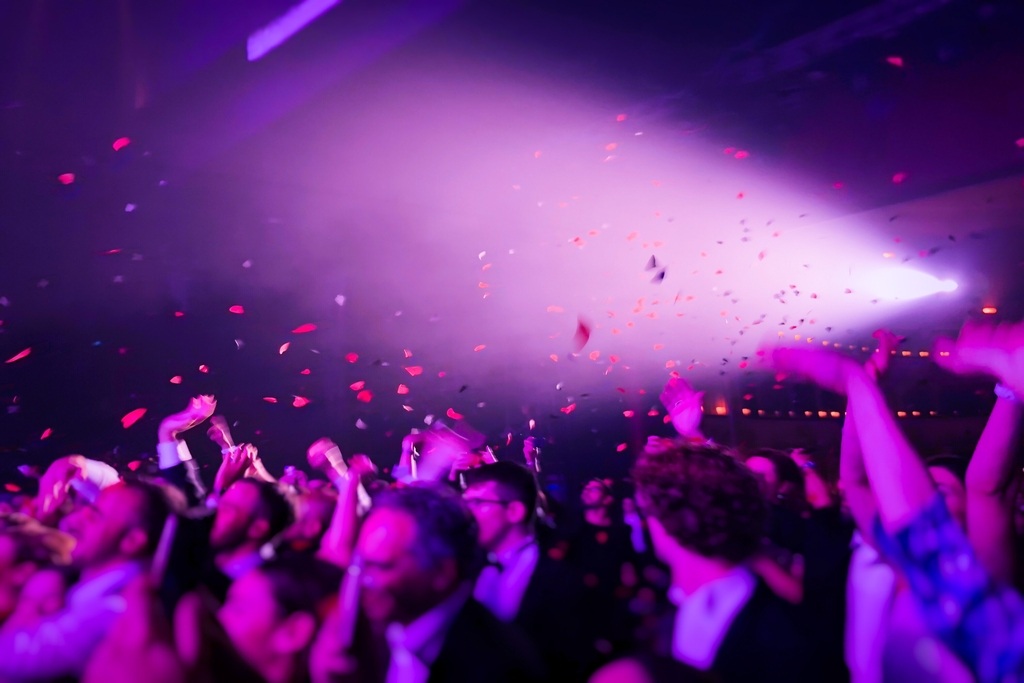 Eine ausgelassen feiernde Menschenmenge auf einer Betriebsfeier, von unten fotografiert. Viele Hände sind in die Luft gestreckt, während rotes Konfetti in einem hellen, lila-pinken Scheinwerferlicht von der Decke regnet. Die Atmosphäre ist energiegeladen u