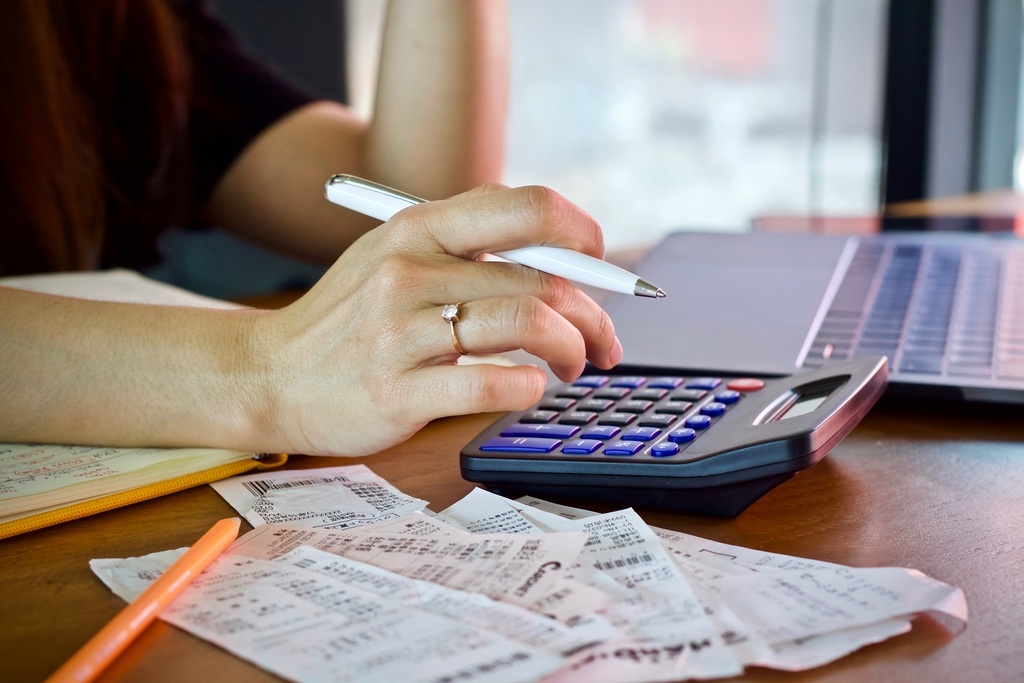Eine Nahaufnahme zeigt eine Person, die an einem Schreibtisch sitzt und ihre Finanzen verwaltet. Vor ihr liegt ein großer Haufen Belege oder Tickets, ihre Hand mit einem Stift schwebt über einem Taschenrechner. Die Szene deutet auf die Berechnung von Ausga