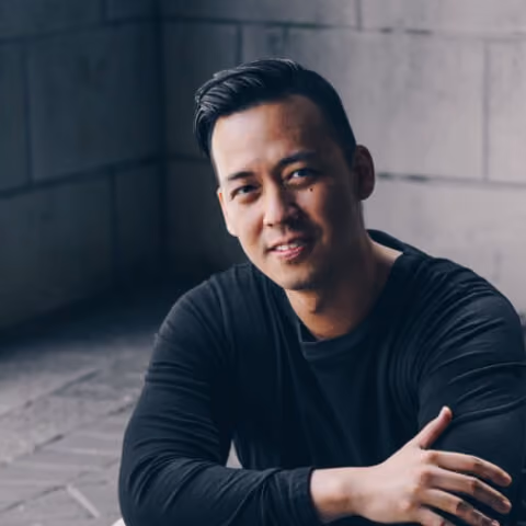Smiling man with short black hair in a black long-sleeve shirt sitting in front of a gray wall.