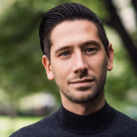 Portrait of a man with short dark hair and blue eyes wearing a black turtleneck outdoors.