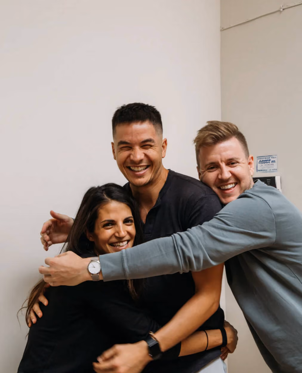 Three friends smiling and hugging each other in a warm group embrace indoors.