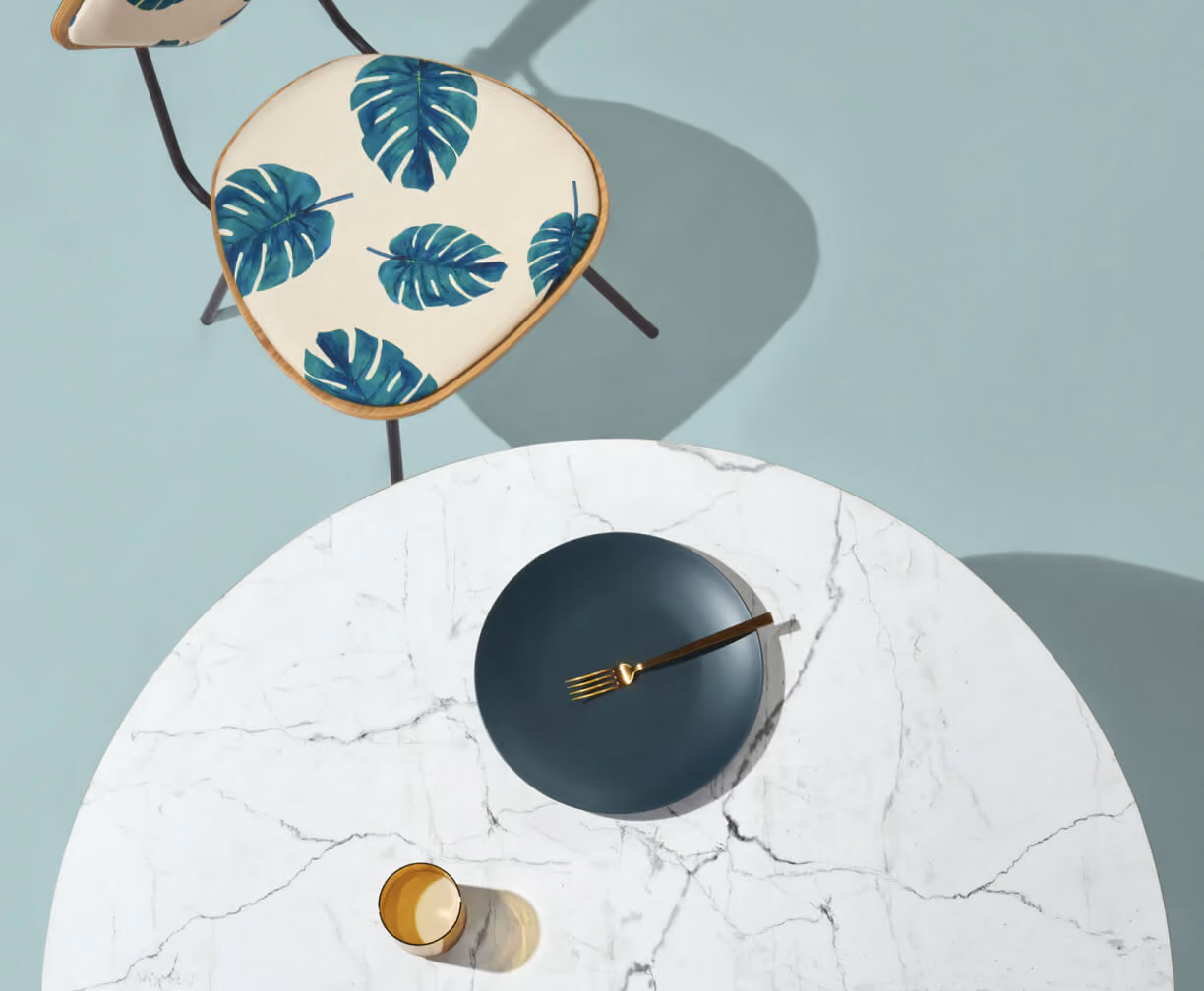 Top view of a marble table with a dark plate, gold fork, gold cup, and a chair with leaf pattern cushion.