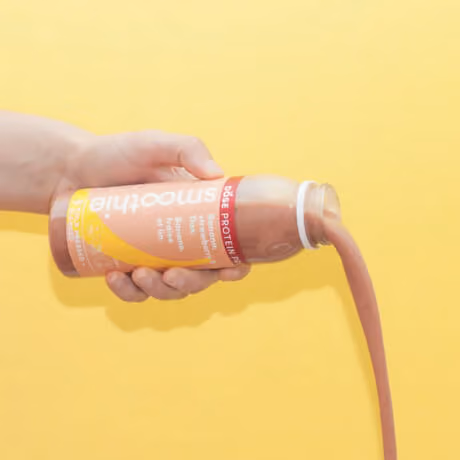 Hand pouring a brown protein smoothie from a bottle against a yellow background.